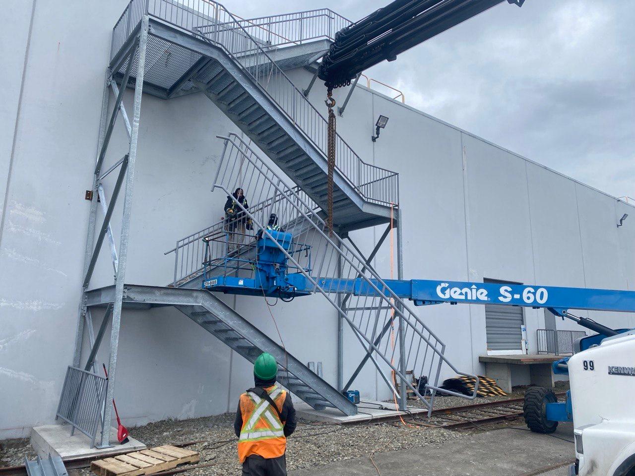 Jeff and Simon Ironworks crew installing a galvanized structural steel egress stair with code-compliant picket guardrails from a boom lift on a Burnaby commercial building